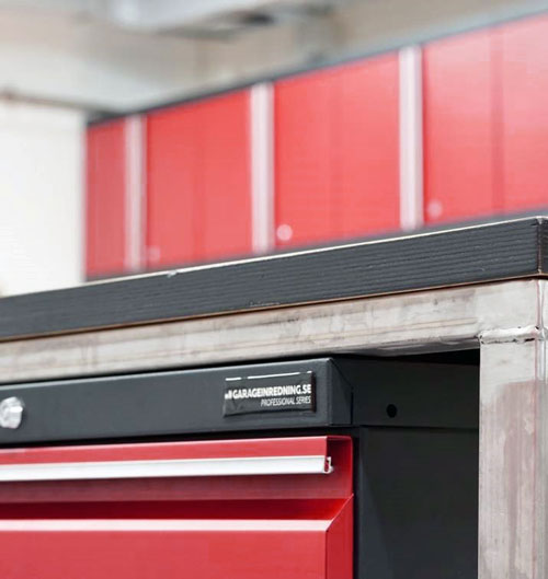 En närbild av ett rött verktygsskåp med en snygg svart bänkskiva visar den djärva logotypen "GARAGEINREDNING.SE PROFESSIONAL SERIES." I bakgrunden är fler röda skåp något suddiga, vilket antyder en organiserad Garageinredning-verkstadsmiljö.