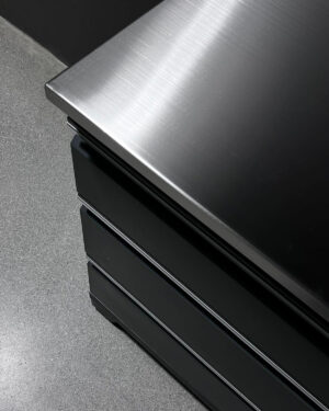 Närbild på den eleganta, moderna Bänkskiva rostfri 71cm PRO SERIES med sin topp i borstat stål. Ytan reflekterar ljus och framhäver dess släta struktur. Det grå golvet ger kontrast till denna glänsande möbel.