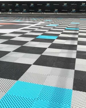 En närbild av ett flerfärgat rutigt golv med nyanser av grått, svart, vitt och aquablått har SWISSTRAX SMOOTHTRAX GOLVPLATTOR i Royal-Blue. En svart bakgrund visar ett upprepande logotypmönster.