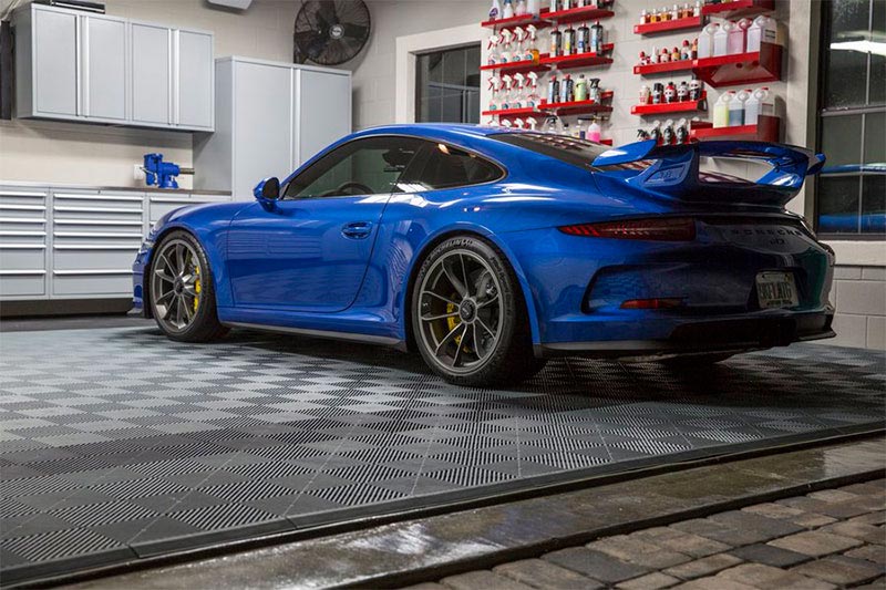 En blå sportbil står parkerad i ett välorganiserat garage med klinkergolv. Hyllor fyllda med bilvårdsprodukter är monterade på väggen och ett verktygsskåp syns i bakgrunden.