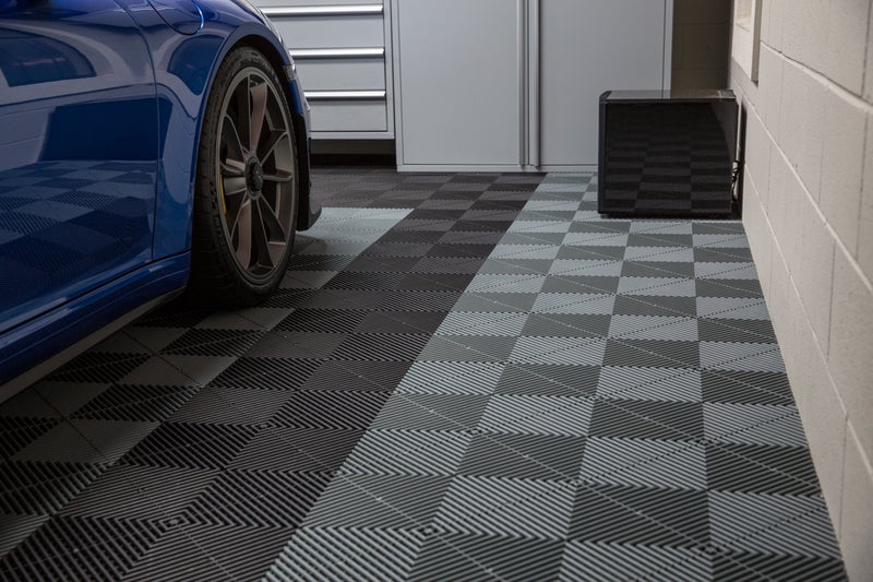 En blå bil är delvis synlig, parkerad på ett garagegolv med ett geometriskt svart och grått kakelmönster. Ett skåp och en svart låda står mot väggen, vilket skapar ett organiserat och modernt utrymme.