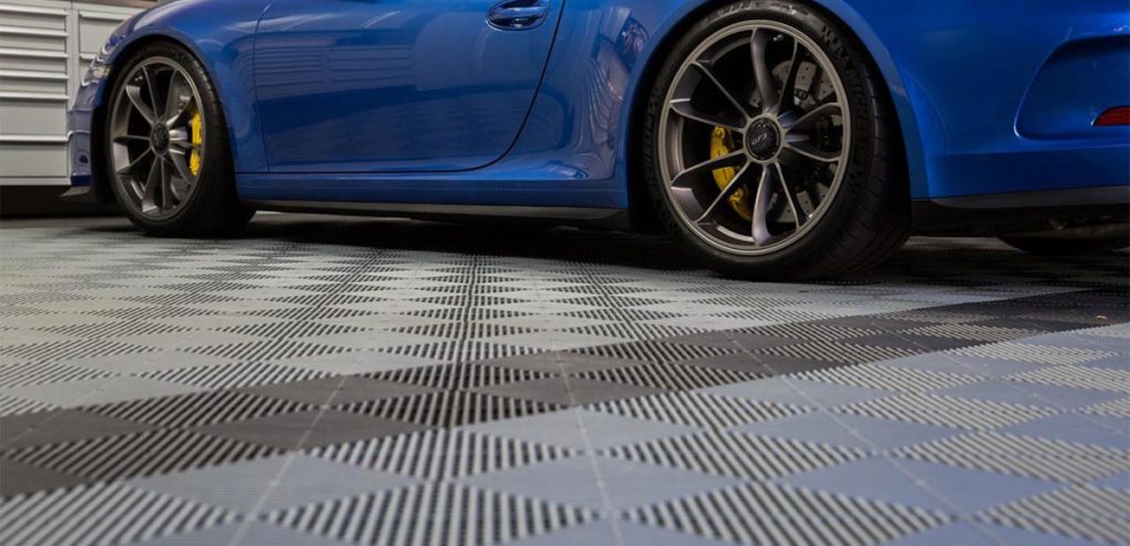 En blå sportbil med gula bromsok står parkerad i ett garage med rutigt grått och svart golv. Garaget har vita skåp i bakgrunden.