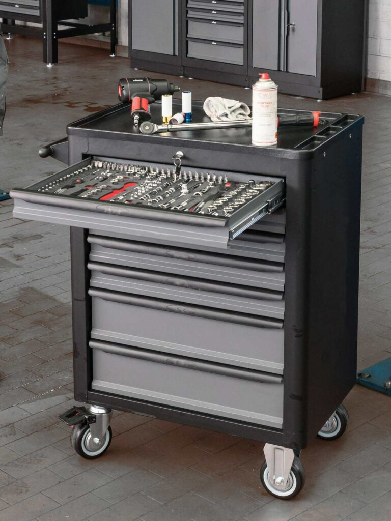 Ett verktygsskåp med hjul, med flera lådor och en öppen låda med olika verktyg, passar sömlöst in i garageinredningen. En burk spray och en trasa vilar ovanpå. Skåpet är placerat i en verkstad tillsammans med andra förvaringsenheter i bakgrunden.
