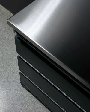 En närbild av ett elegant, metalliskt skiva med en yta av borstad stål, placerad på en slät, stengrå garagebänk. Skåpet har tre lådor staplade prydligt ovanför varandra.