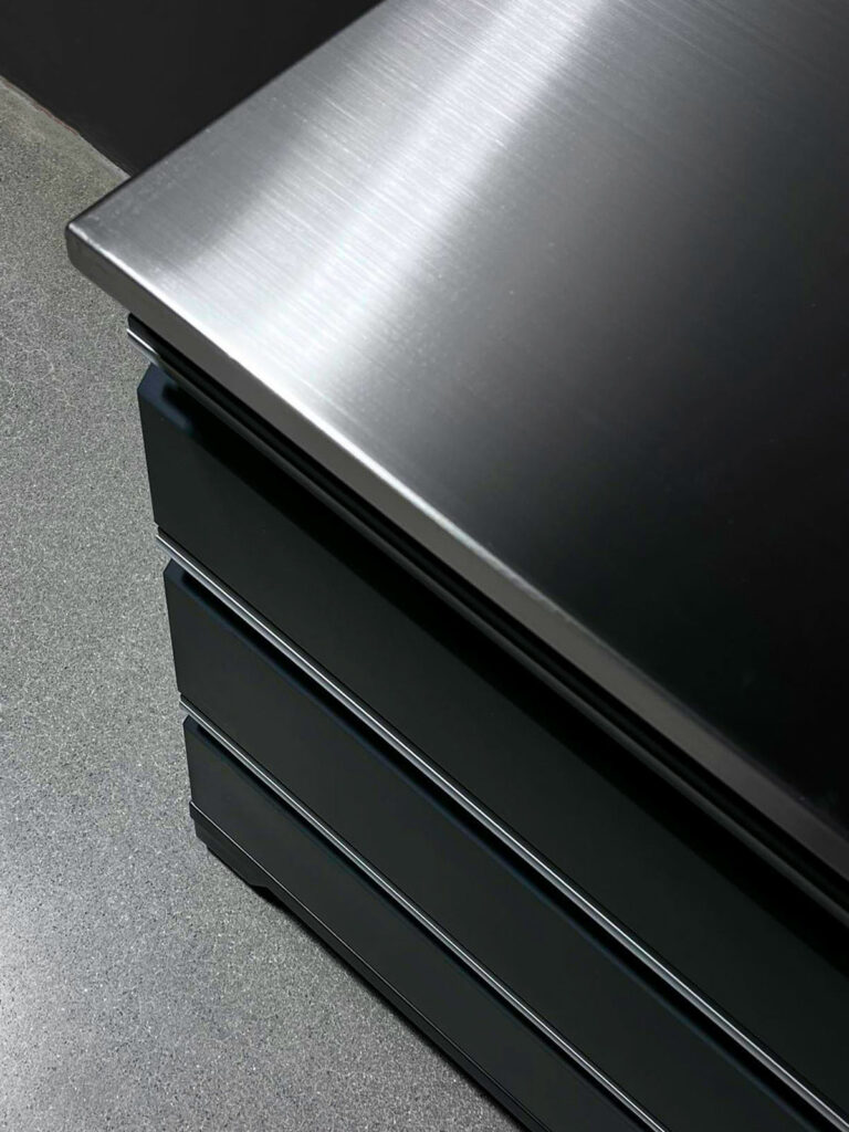 En närbild av ett elegant, metalliskt skiva med en yta av borstad stål, placerad på en slät, stengrå garagebänk. Skåpet har tre lådor staplade prydligt ovanför varandra.