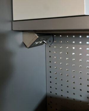 En antracitfärgad Schneider Exxact Hörnbox med trevägsuttag, med metallstruktur och perforerade väggar, omsluter en liten vinklad armatur med synliga kablar. Armaturen är monterad under en hylla i en svagt upplyst industriell eller mekanisk miljö.