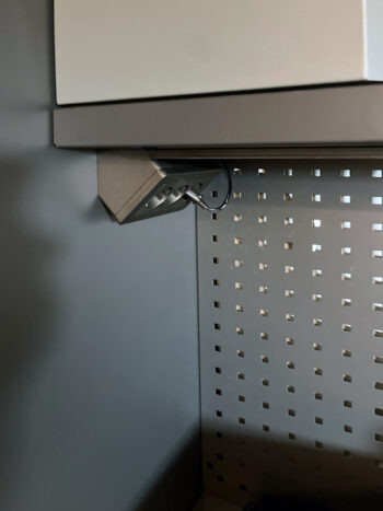 En antracitfärgad Schneider Exxact Hörnbox med trevägsuttag, med metallstruktur och perforerade väggar, omsluter en liten vinklad armatur med synliga kablar. Armaturen är monterad under en hylla i en svagt upplyst industriell eller mekanisk miljö.